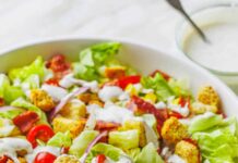 Close up of homemade garden salad in a large white bowl topped with croutons and drizzled with Ranch dressing, with two serving spoons.