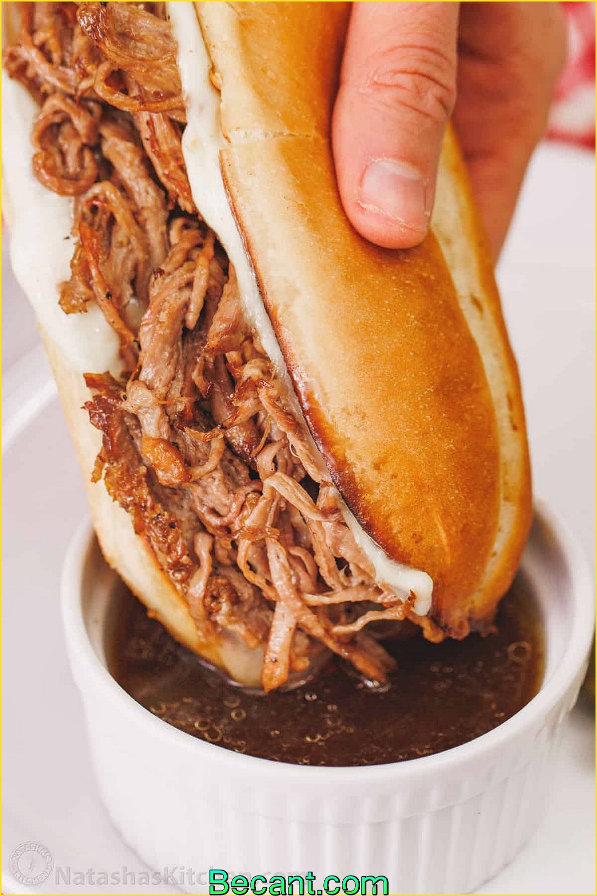 Une main trempe un sandwich au bœuf râpé dans un plat allant au four avec de la graisse de bœuf.