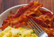Fluffy scrambled eggs on a plate garnished with chives, next to crispy bacon and a fork.